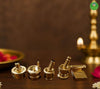 Four brass oil lamp wicks and wick holder components arranged on a dark wooden surface, with a softly blurred brass diya and pink flower petals in the background.